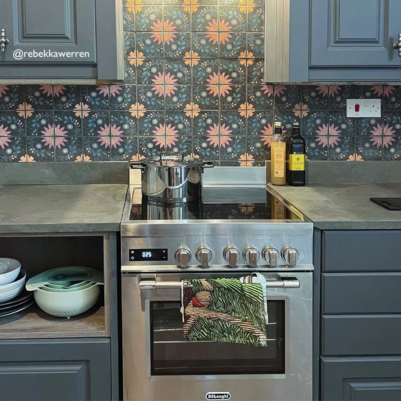 Stainless steel de’longhi electric range with six control knobs and digital display shown with kelmscott blackwell patterned tile background