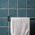 A plush white towel hangs on a dark metal rack against textured teal lagoon field tile wall