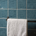 A plush white towel hanging on a dark metal rack against teal lagoon field tile wall background