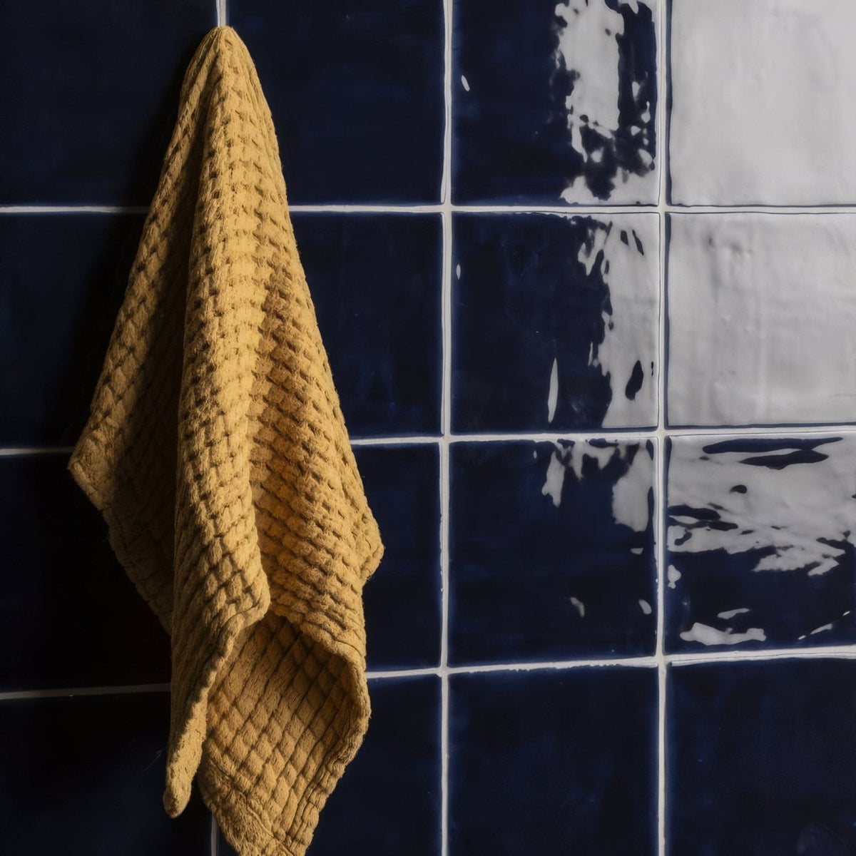 A mustard-yellow textured towel with a woven pattern hanging against dark blue lapis field tile wall