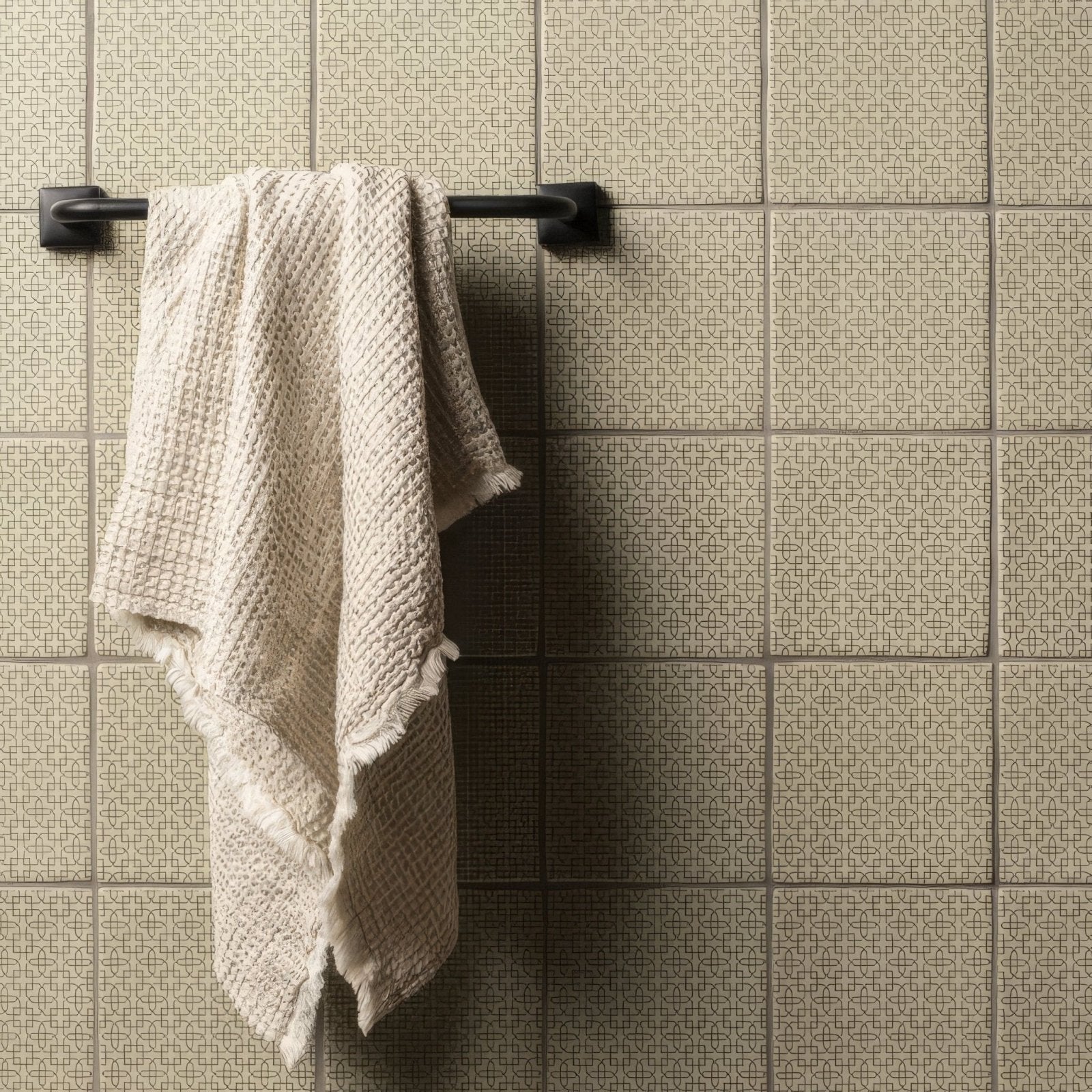 A soft off-white waffle-weave Manoir Maroc pumice towel with frayed edges hanging on a black metal rack