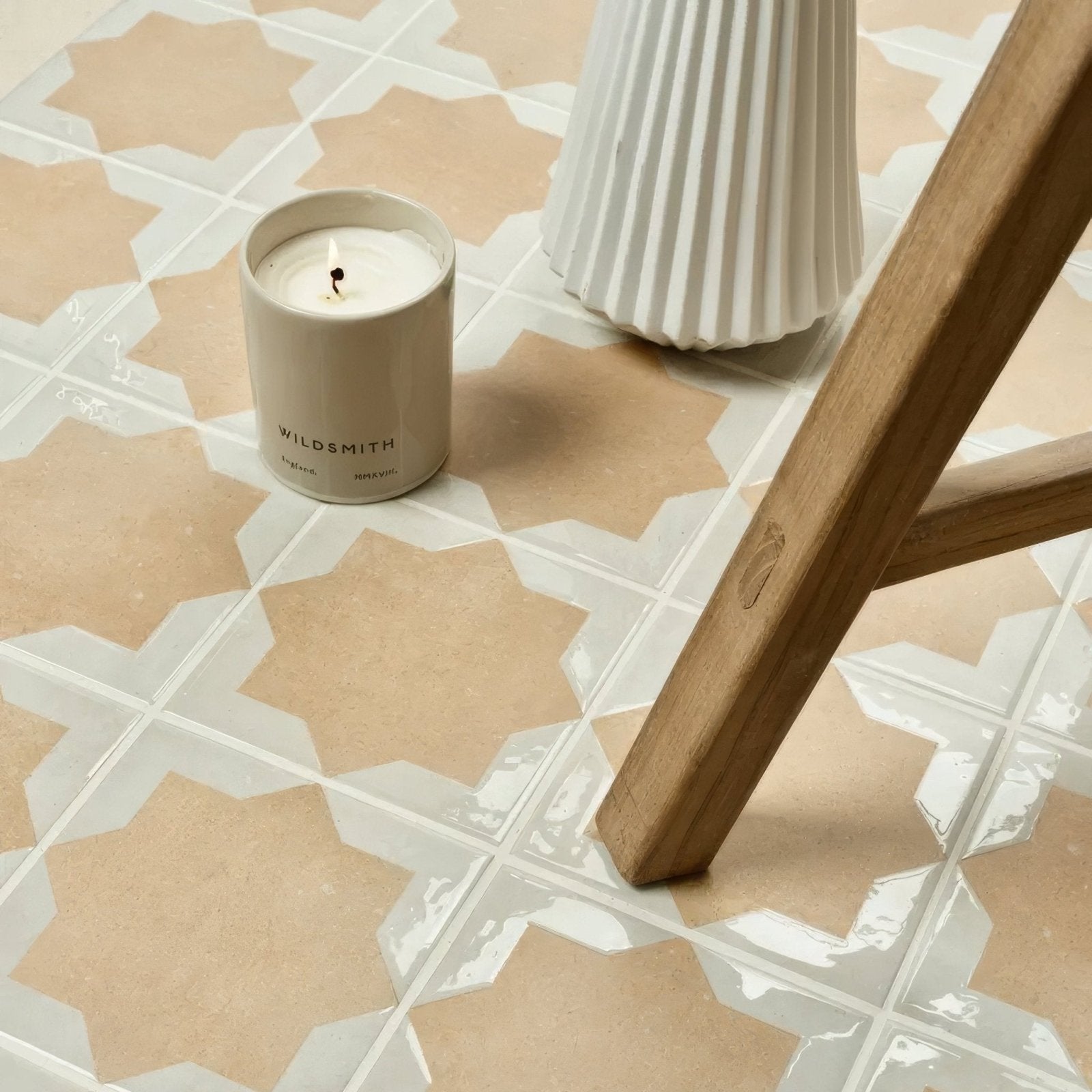 A beige ceramic candle jar with a minimalist label and single wick resting on patterned tiles by menara cotto star porcelain