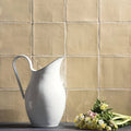A sleek white ceramic pitcher with a curved metallic handle stands against warm beige mimosa field tile backdrop