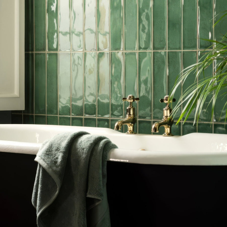 Vintage-style brass faucet set with cross handles mounted on white clawfoot bathtub against glossy montblanc emerald tiles