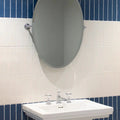 Oval-shaped chrome-framed bathroom mirror mounted above white ceramic sink with matching faucet on nina campbell rivage ivory glazed wall tile