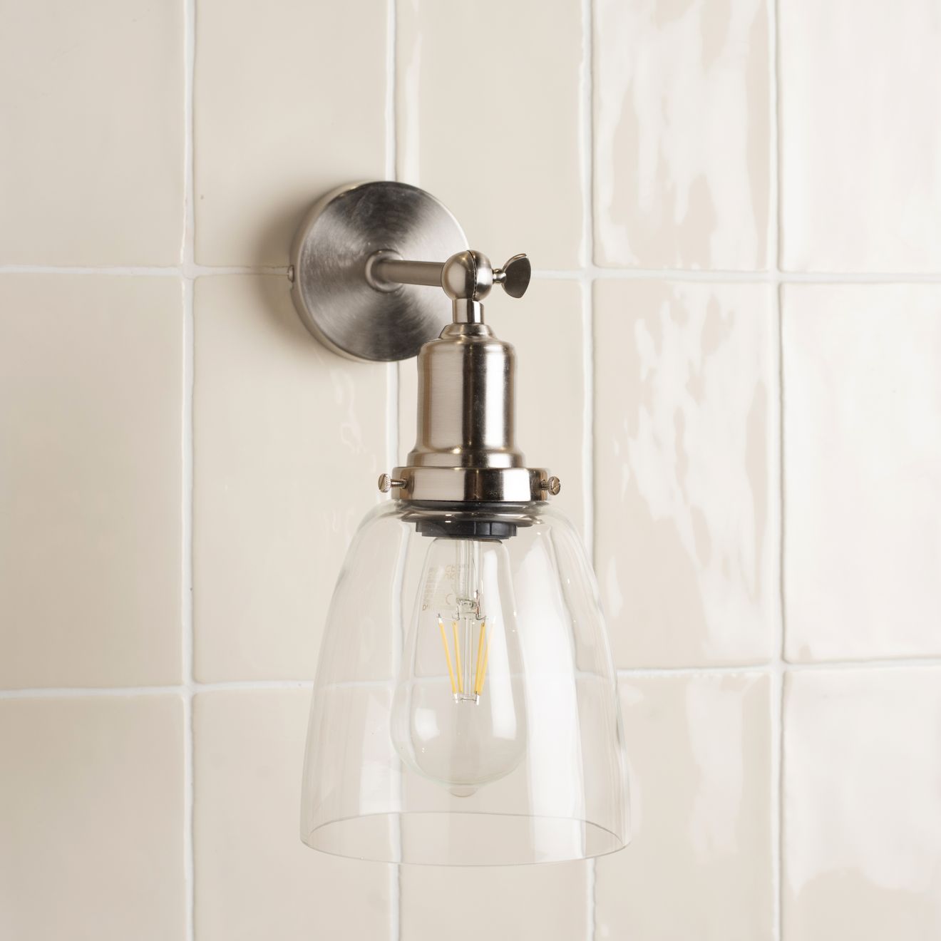 A sleek brushed nickel wall sconce with a clear glass dome and exposed filament bulb in palomino brick tile setting