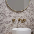 Gold-finished wall-mounted faucet with cross handles and curved spout against patterned eastern mist pine single tile wallpaper