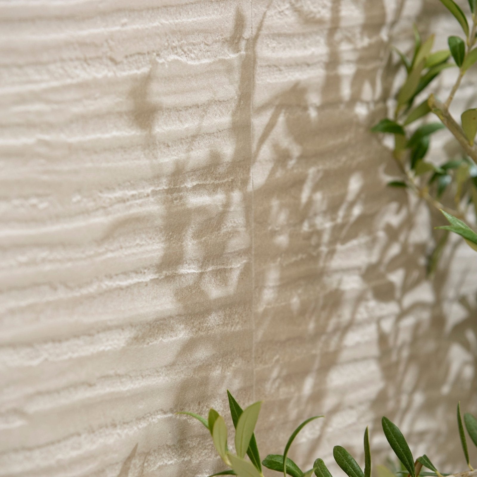 Textured off-white porcelain mink wall with subtle horizontal ridges and soft shadows from green foliage by plastercraft linea mano