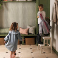 A child in a light blue denim dress with lace collar and pink boots stands near a green bench with patterned pillows in promenade hastings 60x30cm