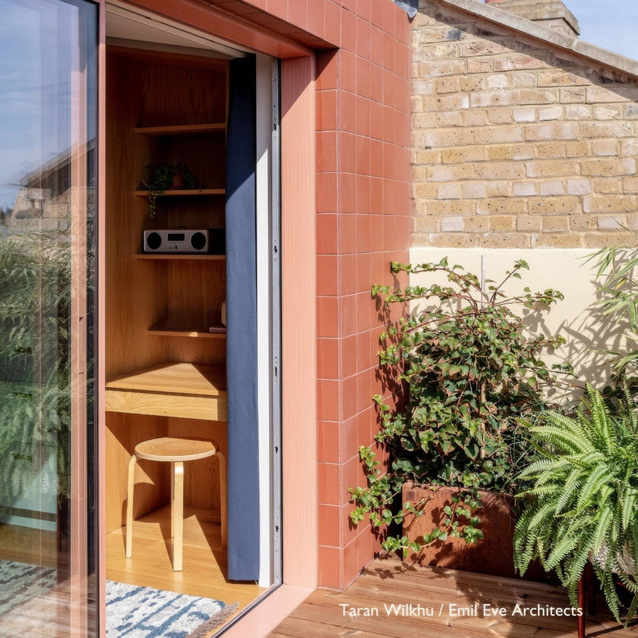 A cozy sunlit corner office featuring quarry red square terracotta-tiled walls with a wooden desk and stool