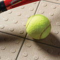 A bright yellow tennis ball with distinct brown seams resting on textured light beige quartz multidisc quarry tile 148x148x9mm