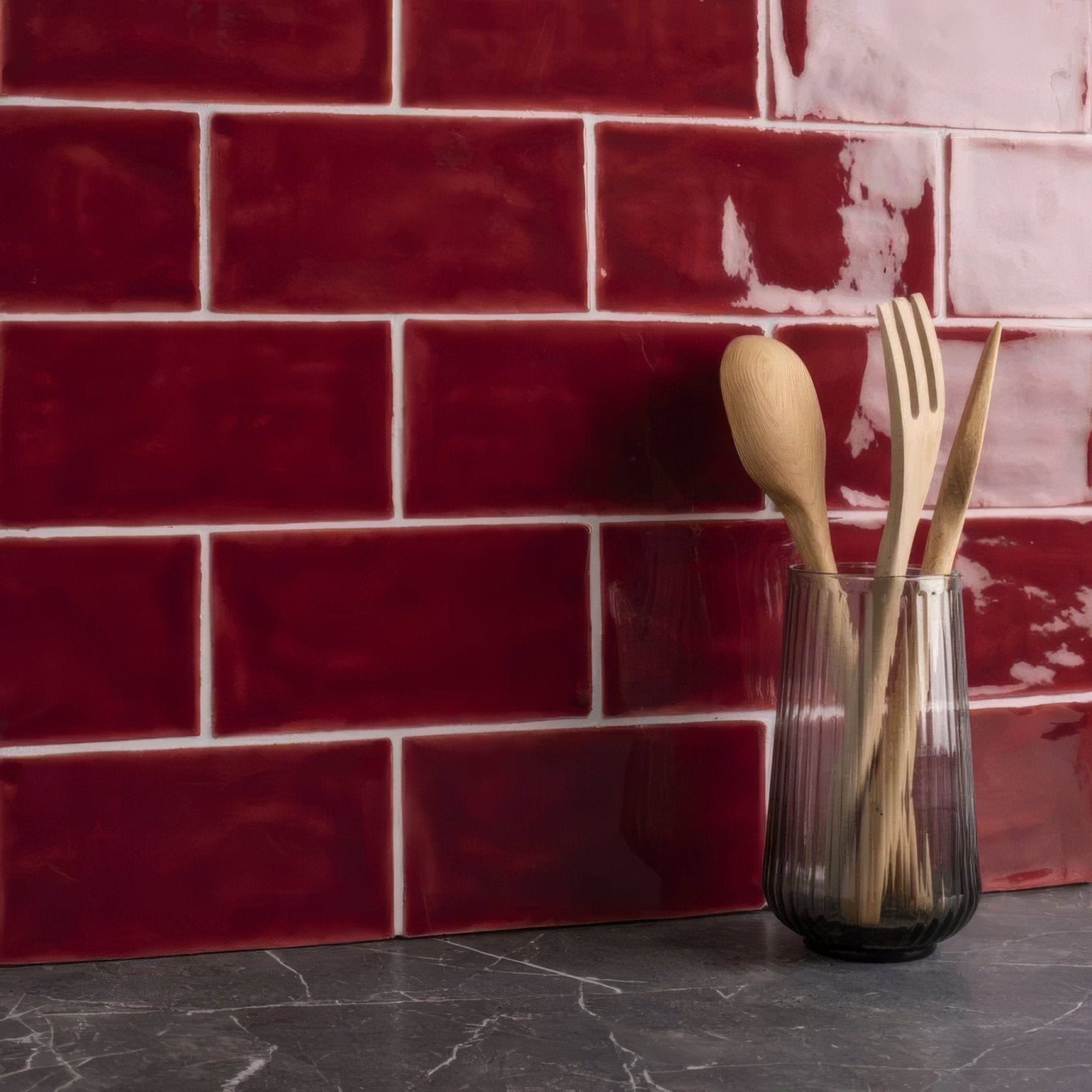 A clear ribbed glass vase holding wooden spoon and fork on a rioja brick tile surface