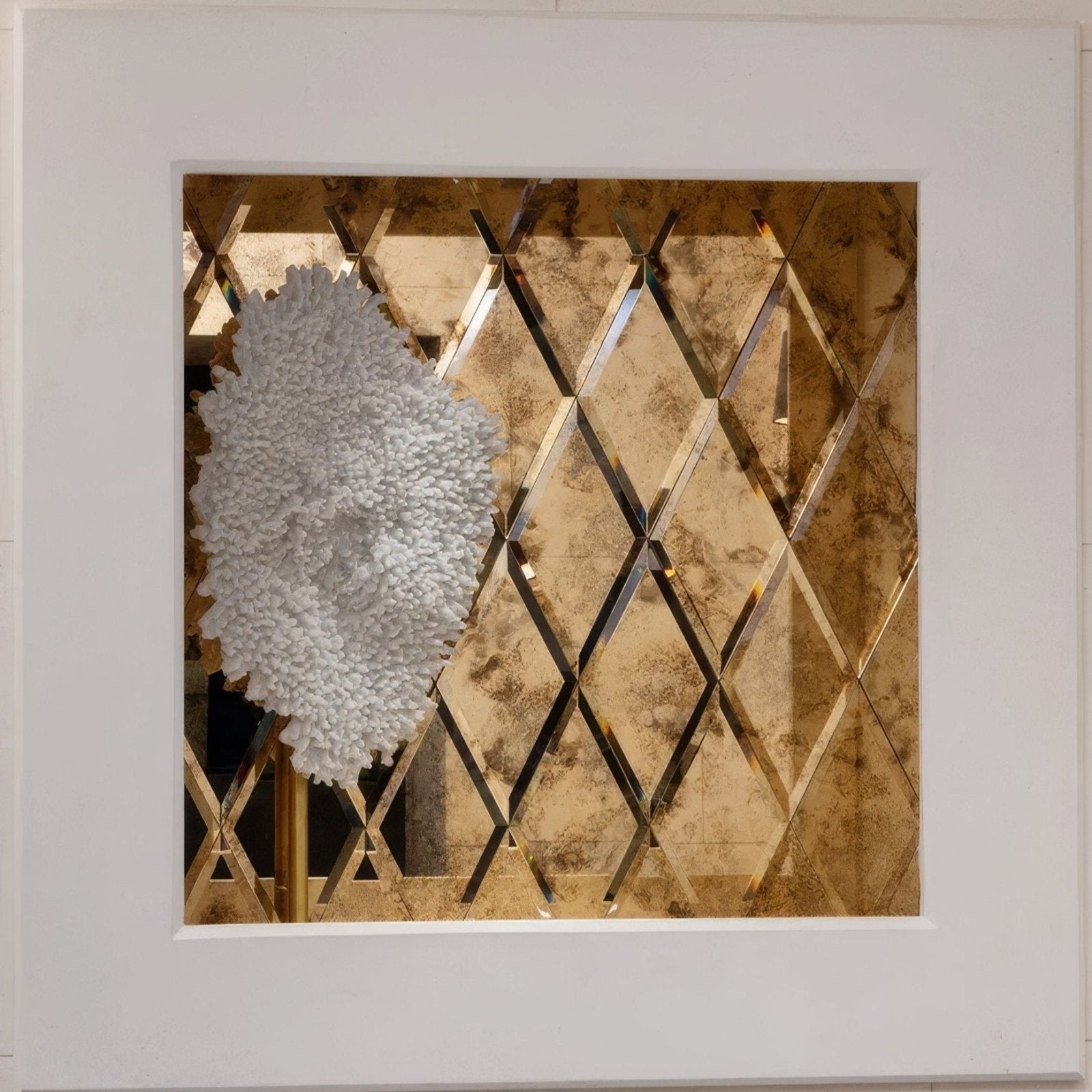 A textured white sculptural wall piece mounted on a reflective gold diamond-patterned glass brass rocco mirror background
