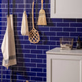 Beige apron with subtle black embroidered logo hanging against vibrant royal blue large brick tiled wall