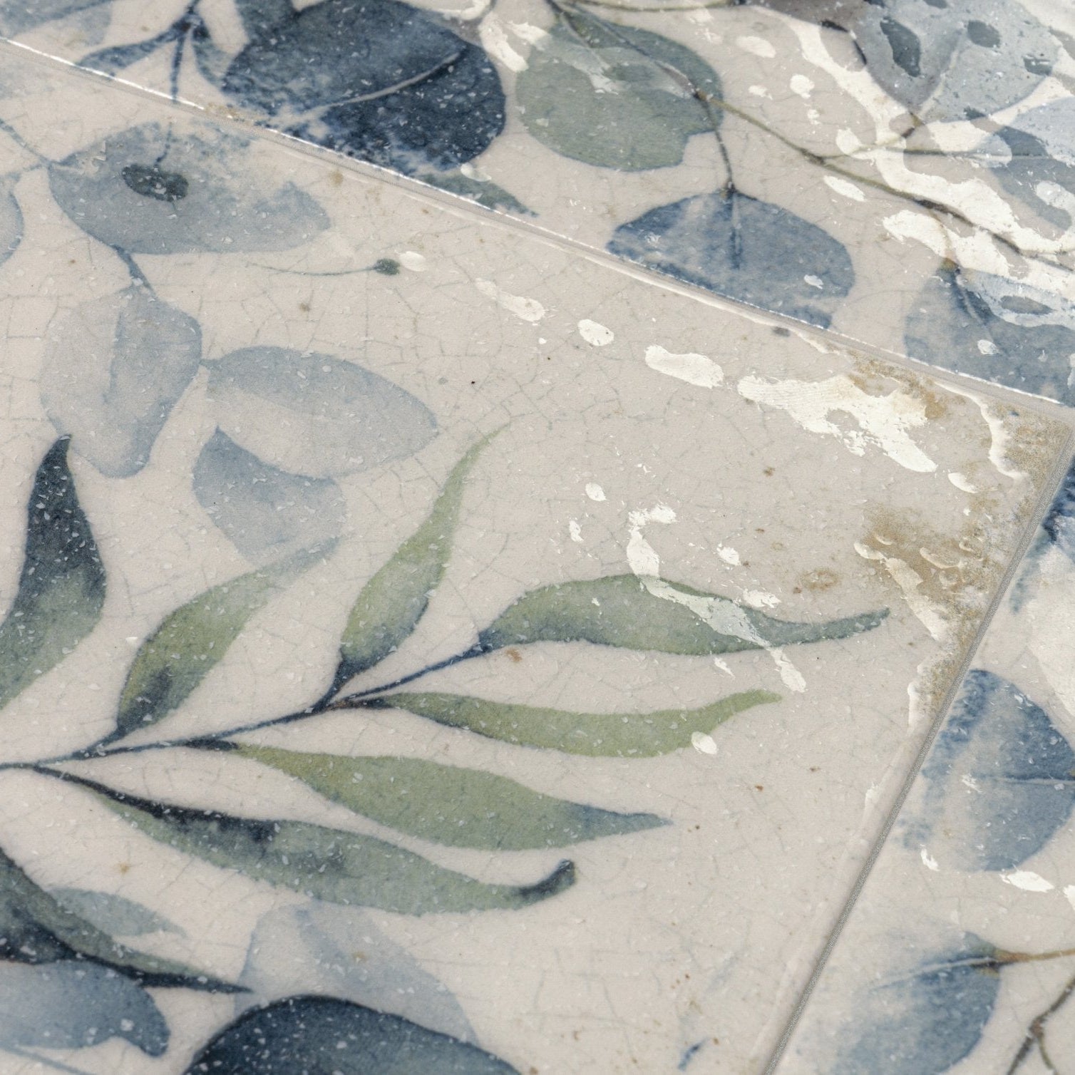 A square sakura delft ceramic tile with crackled glaze and painted green and blue eucalyptus leaves on off-white background