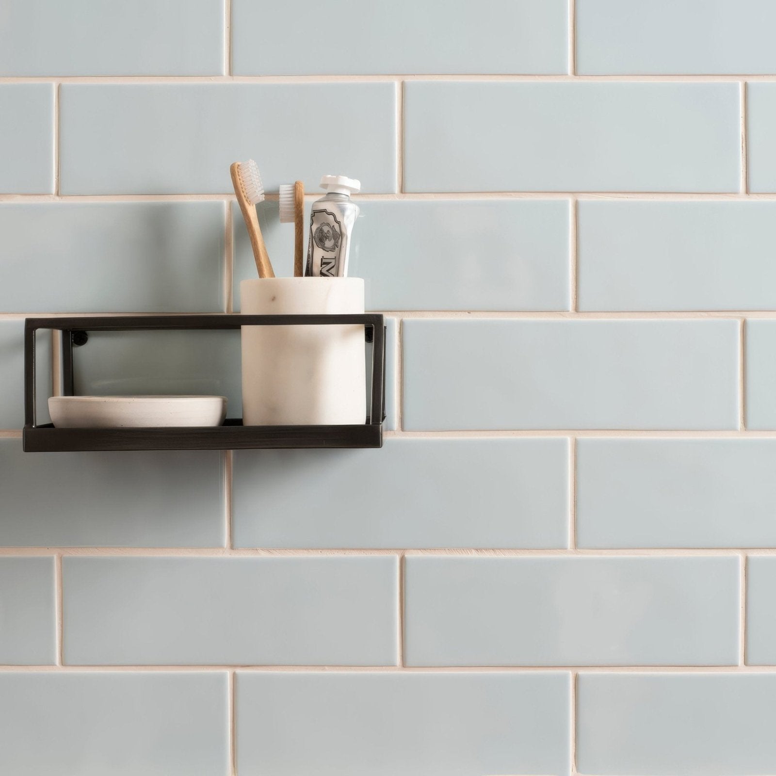 Black metal wall shelf with white ceramic cup and small bowl holding toothbrushes and toothpaste brick tile southwold