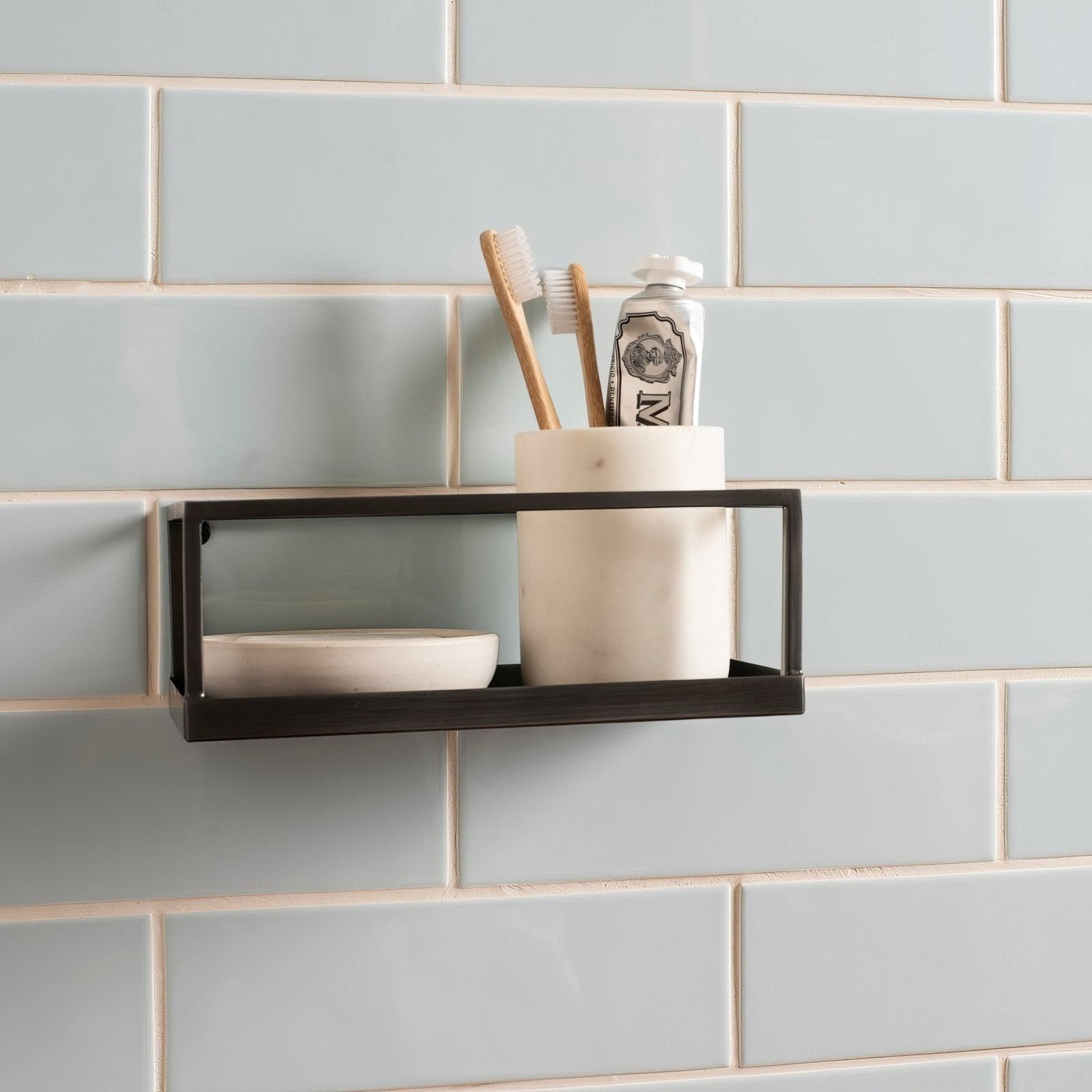 Black metal wall-mounted shelf with rectangular frame holding ceramic toothbrush holder with brushes and toothpaste tube