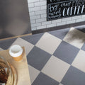A disposable coffee cup with white lid and orange sleeve on a wooden surface next to steel grey flat quarry tile 300x300x9mm