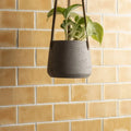 Gray textured concrete hanging planter with dark brown rope straps displayed with straw half tile wall tiles