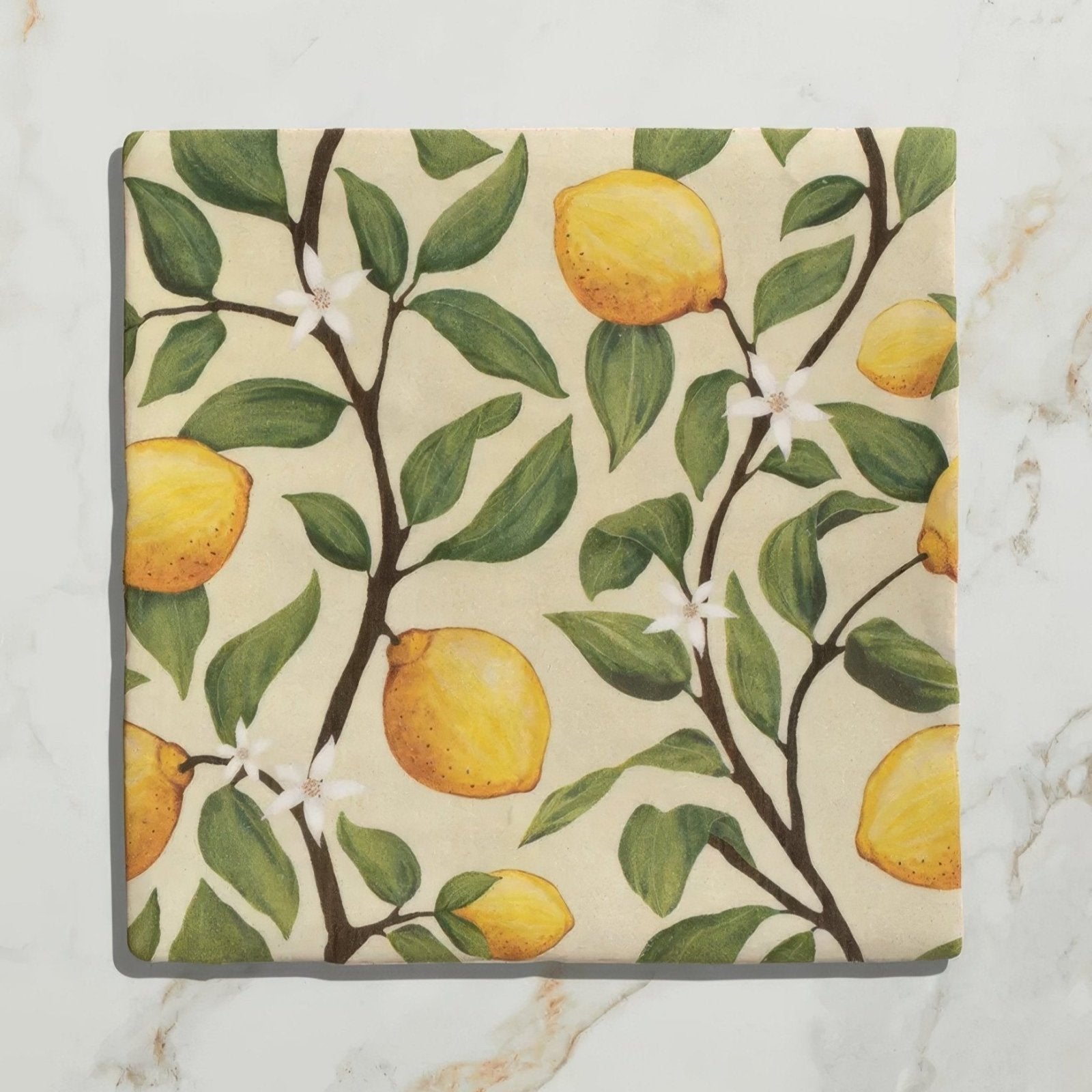 Square ceramic coaster from studio nonna’s kitchen with hand-painted yellow lemons green leaves and white blossoms on off-white