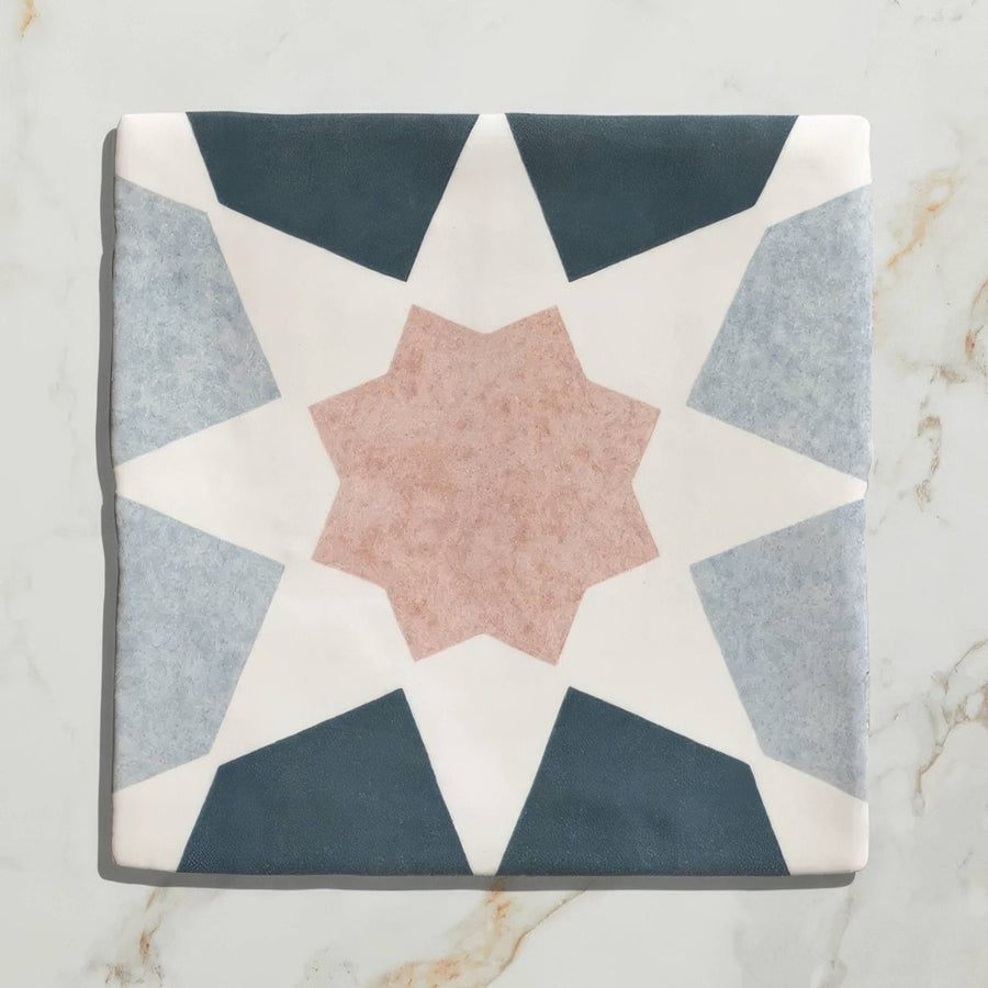 A square ceramic tile from Studio Souk featuring a geometric star pattern in navy gray and blush pink tones