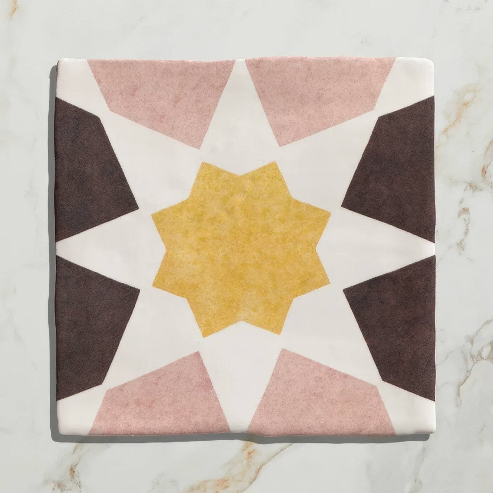 A square ceramic tile with a geometric starburst pattern in muted pink dark brown and mustard yellow tones from Studio Souk