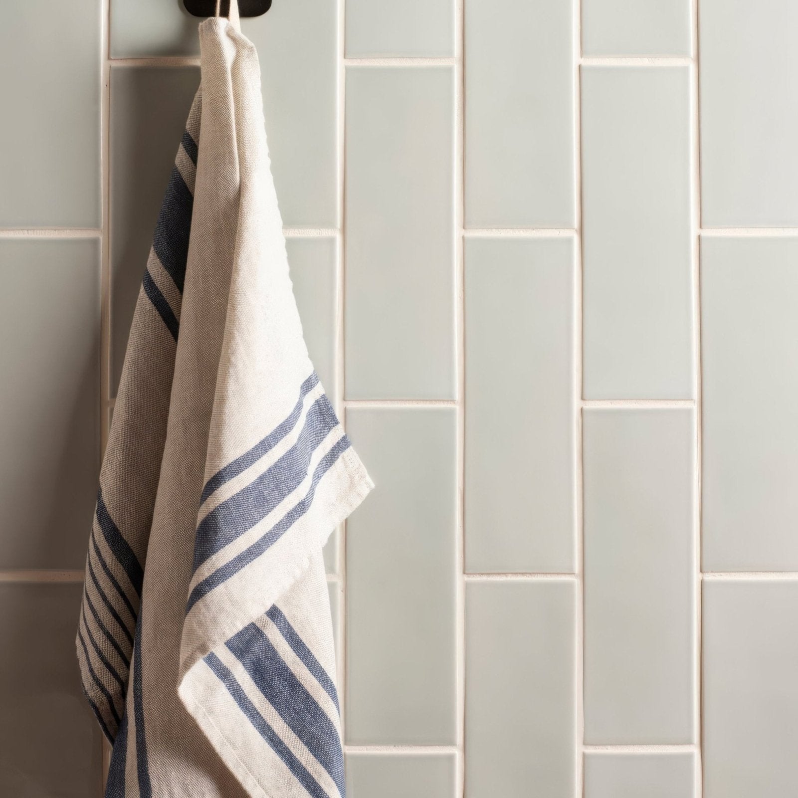 A cream-colored linen towel with navy blue stripes hanging on a hook against light gray brick tile sudbury walls