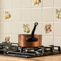 Copper saucepan with black handle on gas stove alongside summer fruits lemon handpainted tile background