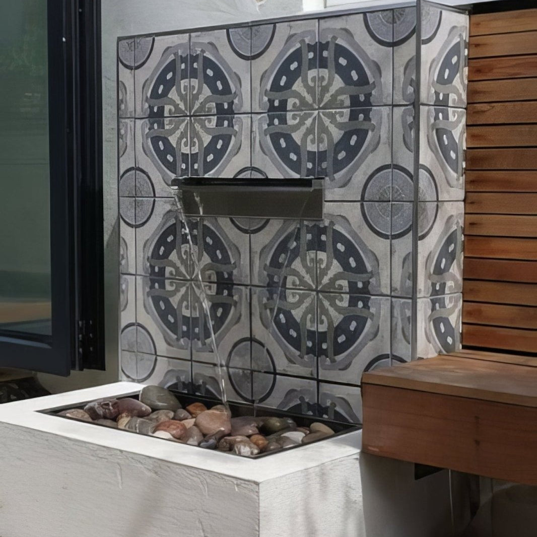 A modern outdoor fountain with a sleek stainless steel spout pouring water into a white concrete basin with river rocks featuring urban aryz urban patterned porcelain tile