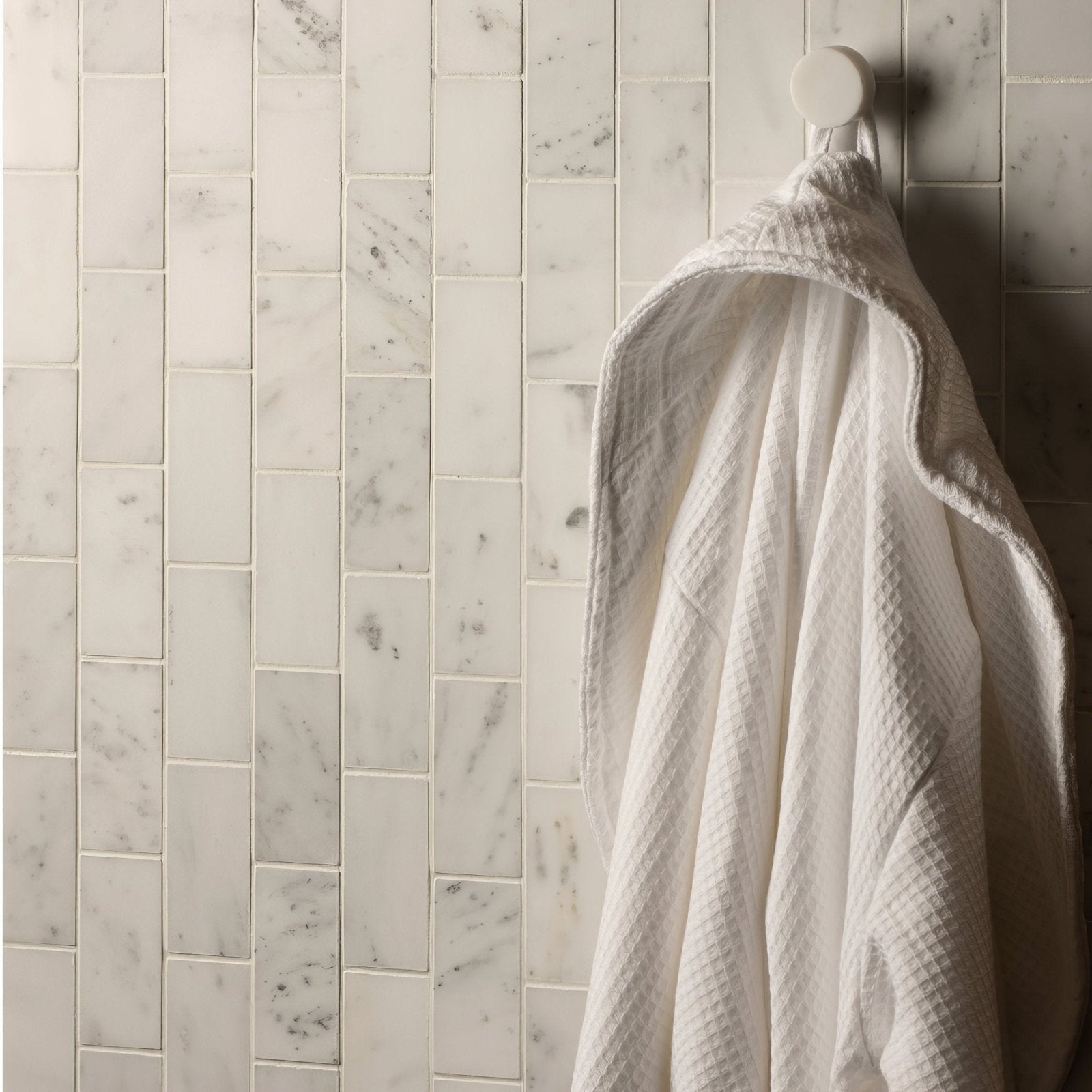 White waffle-weave bathrobe hanging on hook against viano white honed brickbond mosaic marble-tiled wall
