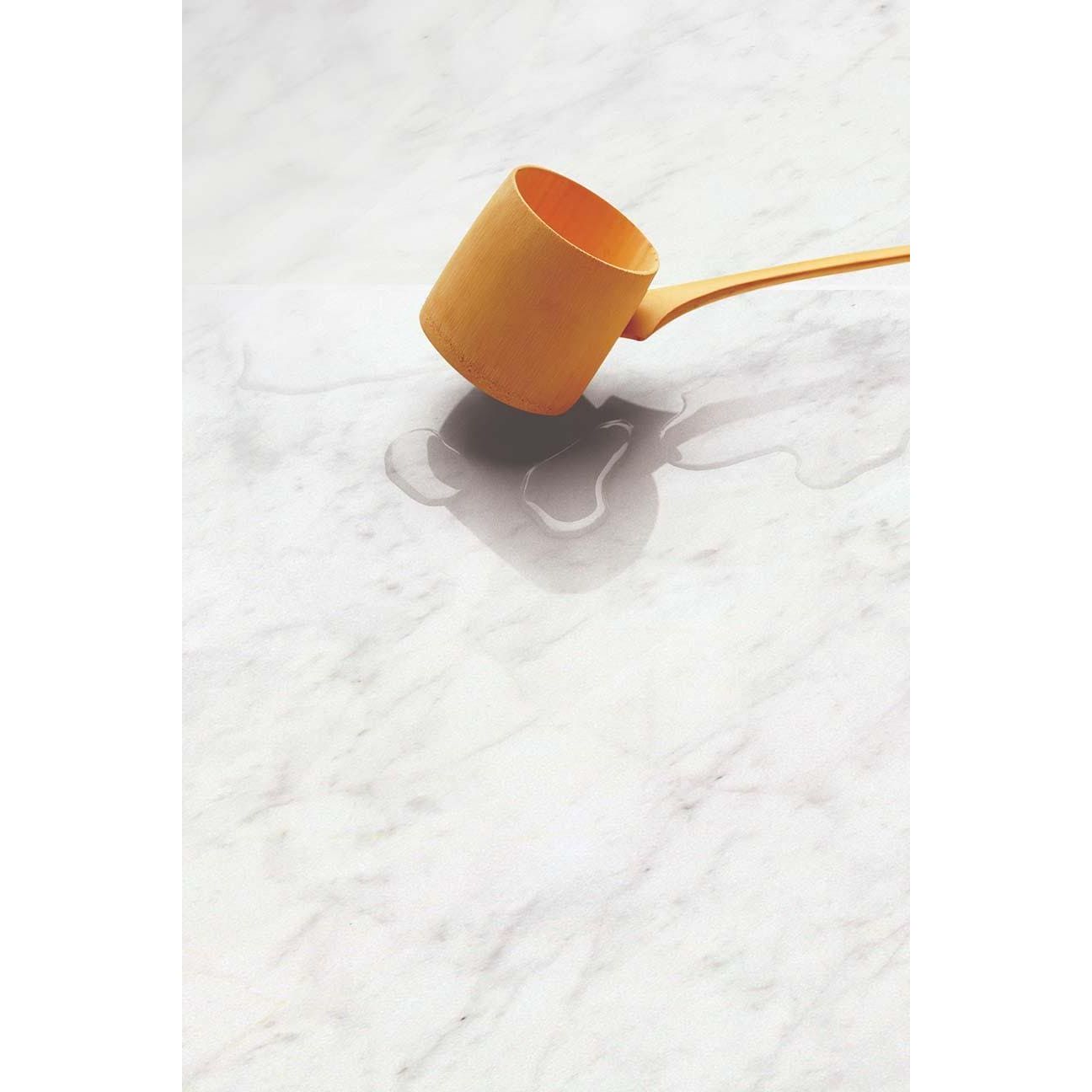 A sleek mustard yellow wooden ladle with a smooth finish on a Python Carrara Porcelain Gloss marble surface